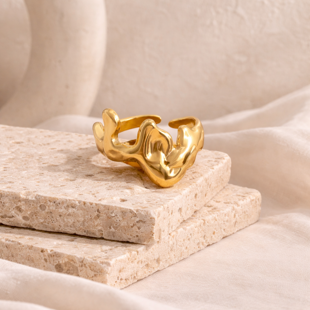 Gold-tone wave-inspired sculpture ring resting on textured stone tiles, showcasing a modern, artistic design from the rings collection in a soft, neutral setting.