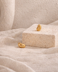 Minimalist gold-toned earrings with a smooth pebble shape, displayed on a natural stone block against a soft, neutral fabric background, part of the Earrings collection.