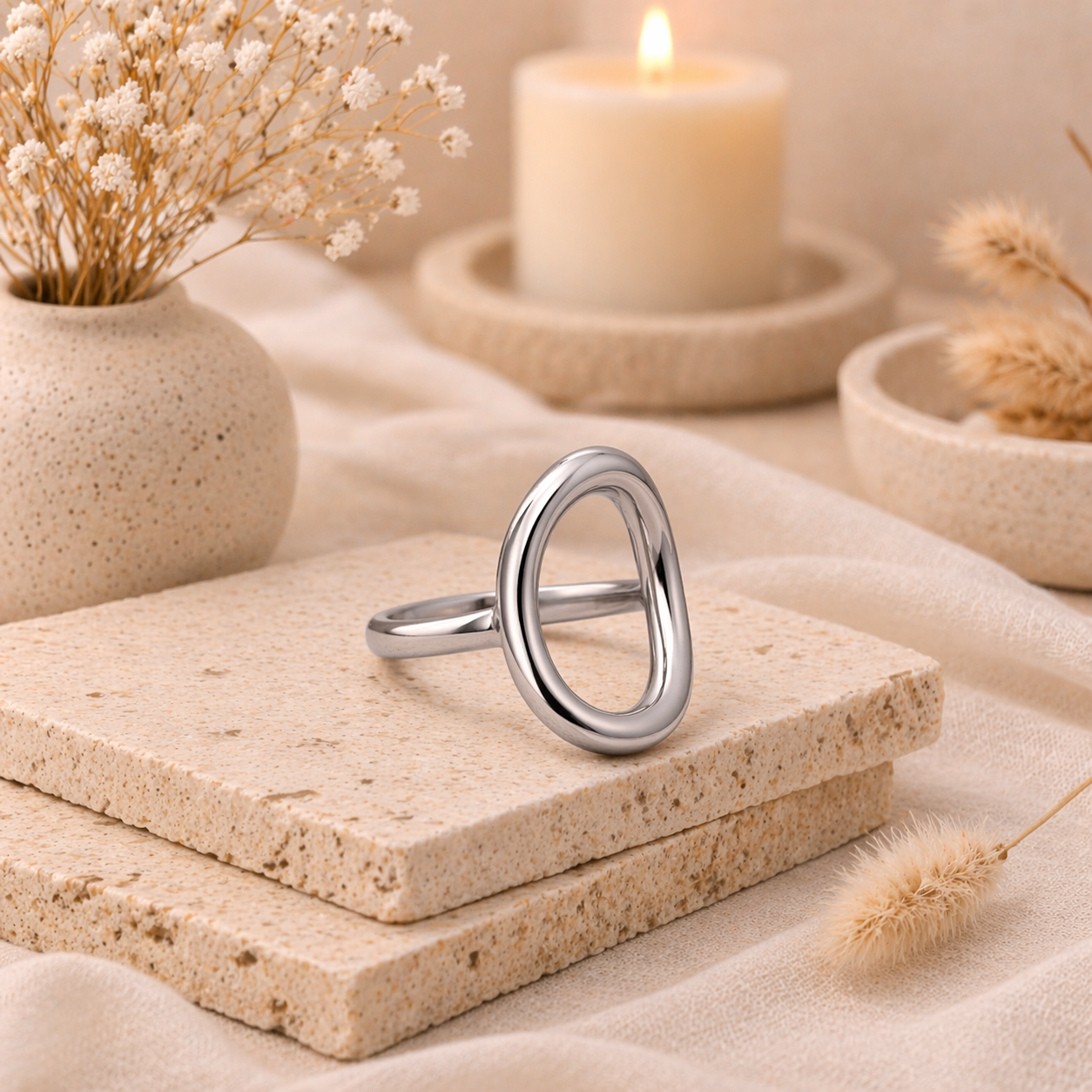 Silver oval-shaped ring from the Silver Collection displayed on textured stone slabs, styled with dried flowers and a softly glowing candle in the background.