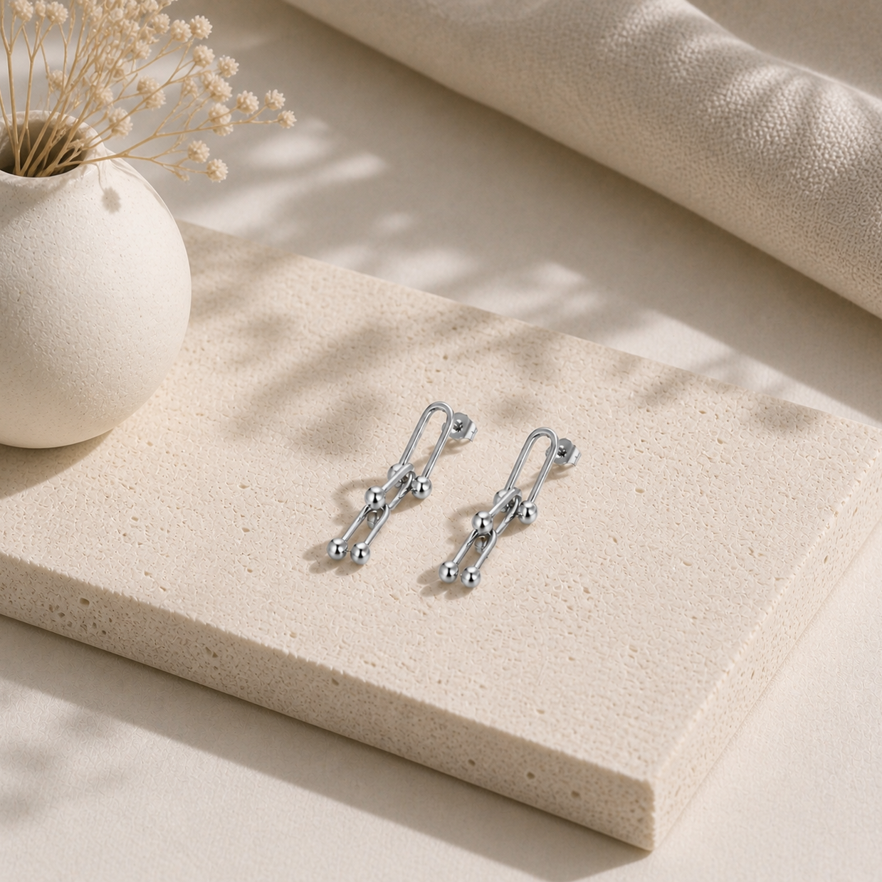 Delicate silver link drop earrings with smooth rounded accents, styled on a textured cream stone alongside minimalist decor, part of the Silver Collection in earrings.