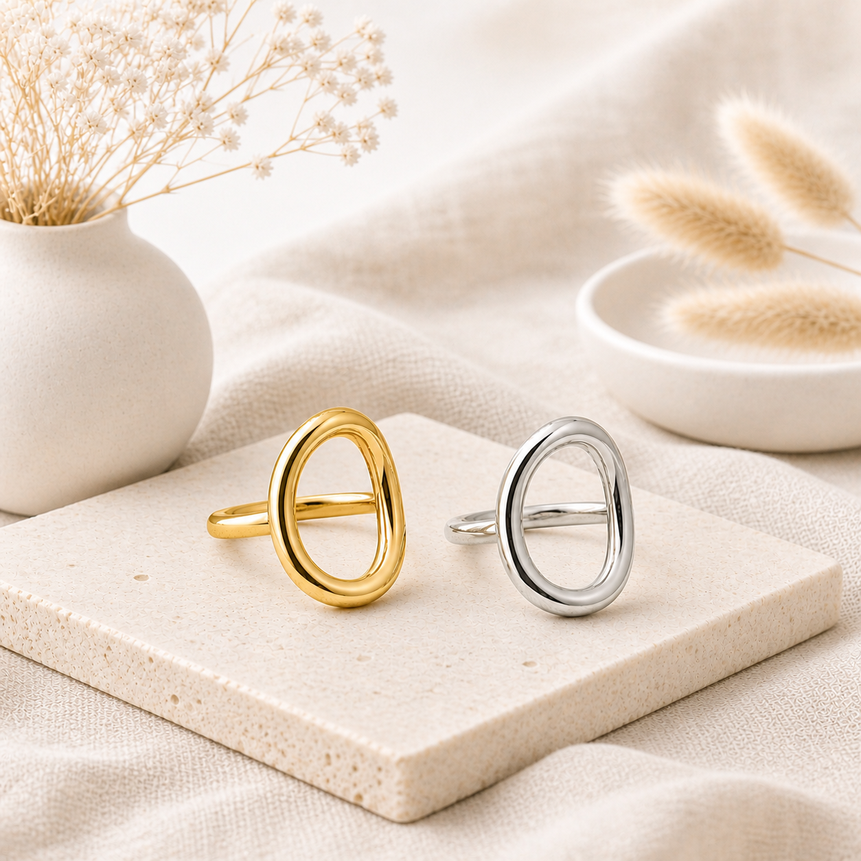 Two oval-shaped rings, one gold and one silver, displayed on a textured stone slab with soft neutral fabric and dried floral accents, part of a silver collection in rings.