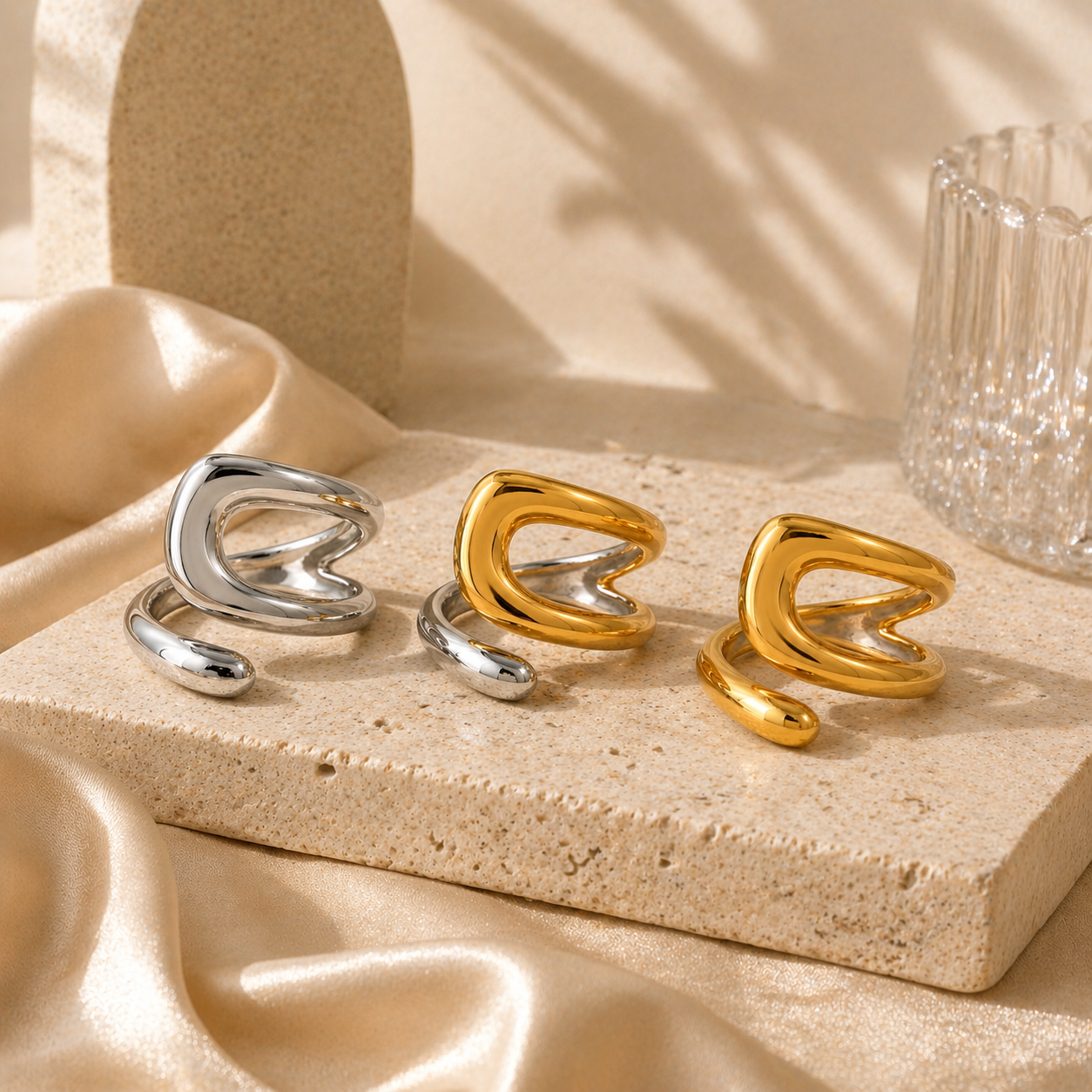 Three modern open rings in polished silver and gold tones displayed on textured stone with soft beige silk and delicate glassware, part of a stylish silver collection.