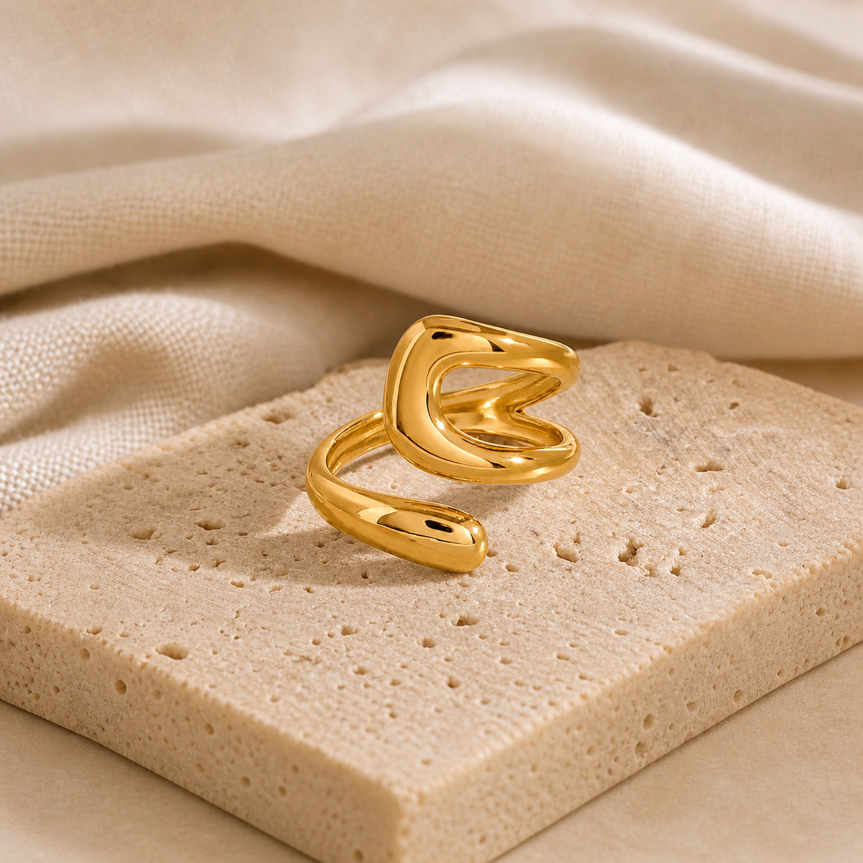 Close-up of a modern, gold-tone open ring with a smooth, sculptural design from the Rings collection, displayed on a textured beige surface with soft fabric background.