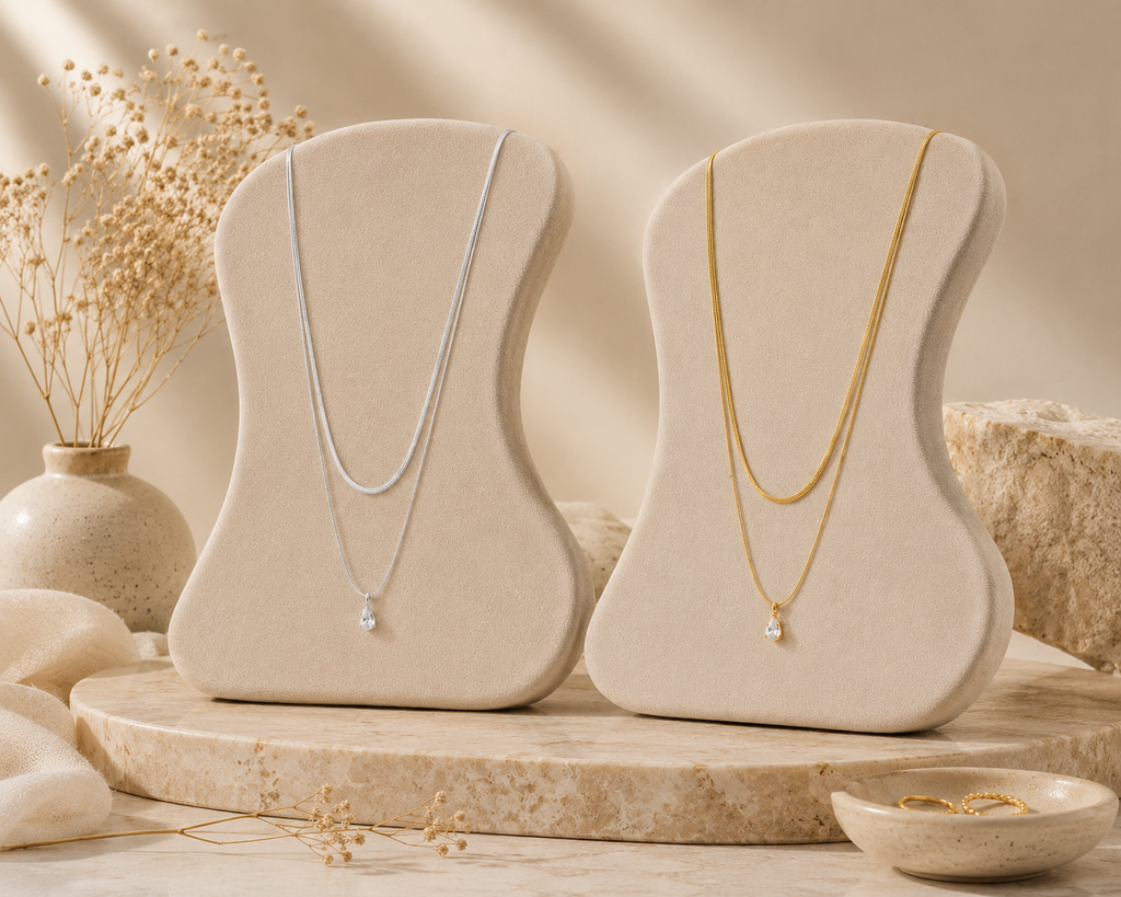 Two elegant layered necklaces displayed on beige velvet stands, one silver and one gold-toned, featuring delicate raindrop crystal pendants from the Silver Collection in a minimalistic setting.